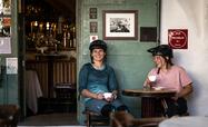 Two women wearing bicycle helmets drink a coffee